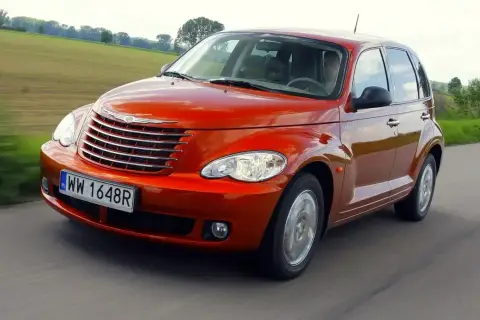 Pomarańczowy Chrysler PT Cruiser jedzie po drodze, za nim zielone pola i drzewa.