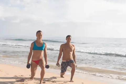 Kobieta i mężczyzna ćwiczą na plaży z hantlami. Ich ruchy to przykład świadomej kinezjologii, poprawiającej siłę i koordynację.
