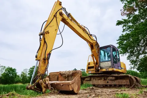 Żółta koparka z pełnym łyżką ziemi, gotowa do pracy. Widać jej gąsienice i mechanizm podnoszenia ramienia, a także zwolnica.