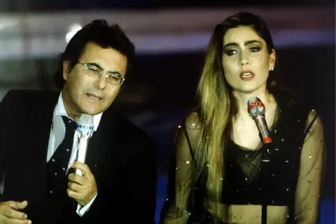 Al Bano i jego nowa partnerka na scenie, w blasku świateł, śpiewają do mikrofonów.