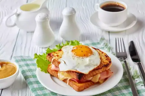 Un délicieux croque monsieur garni d'un œuf au plat, servi avec une tasse de café et une sauce crémeuse.