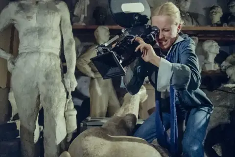 Kobieta z kamerą w pracowni rzeźbiarskiej, jak z filmów Feliksa Falka. W tle antyczne posągi.