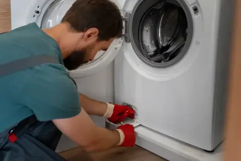 Mężczyzna w czerwonych rękawiczkach otwiera klapkę pralki, by sprawdzić, jak wyczyścić filtr.