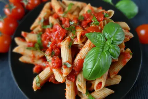 Penne z pomidorami i świeżą bazylią, polane aromatycznym **sosem bazyliowym do makaronu**. Obok gałązka pomidorków koktajlowych.