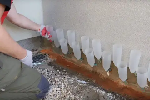 Pracownik w rękawiczkach montuje system do osuszania murów, umieszczając plastikowe rurki wzdłuż ściany.