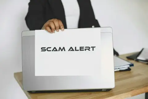 Ostrzeżenie przed oszustwem na Vinted. Ktoś trzyma kartkę z napisem "SCAM ALERT" nad laptopem.