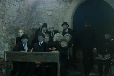 Postacie z "Umarłej klasy" Tadeusza Kantora, w tym uczniowie i nauczyciele, siedzą w ponurej scenerii.
