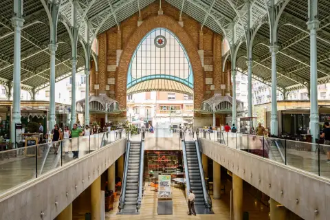 Interior del Mercado de Colón, con sus arcos de hierro y cristal, escaleras mecánicas y puestos de comida.