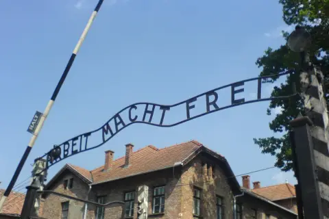 Brama z napisem "Arbeit Macht Frei" nad ceglanymi budynkami pod błękitnym niebem.