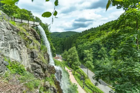 Der Trusetaler Wasserfall stürzt von einem Felsen herab. Ein Weg führt am Wasserfall vorbei, daneben eine Straße und ein bewaldeter Hang.