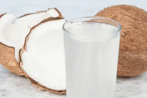 Ein Glas erfrischendes Kokoswasser steht neben einer aufgeschnittenen Kokosnuss. Kokoswasser gesund und lecker!