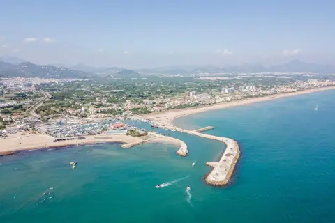Vista aérea del puerto de Oliva con barcos navegando y la playa llena de gente.