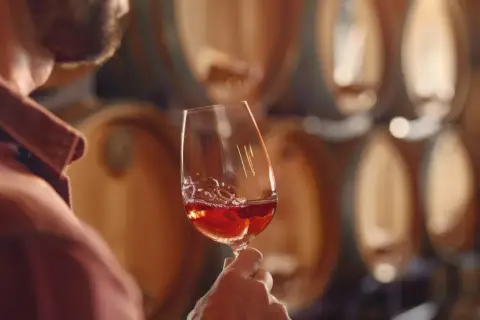 Un uomo assapora un bicchiere di vino rosso nelle cantine di Bolgheri, circondato da botti di legno.