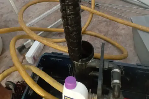 Czarna, brudna rura i żółty wąż wskazują na płukanie instalacji grzewczej. Widać też butelkę z preparatem.