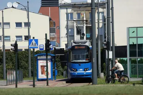 Niebieski tramwaj linii 20 na przystanku, obok rowerzysta i budynki.