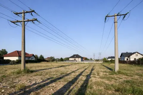 Linia średniego napięcia biegnie nad niezabudowaną działką, z domami w oddali. Widać wyraźną odległość budynku od linii średniego napięcia.