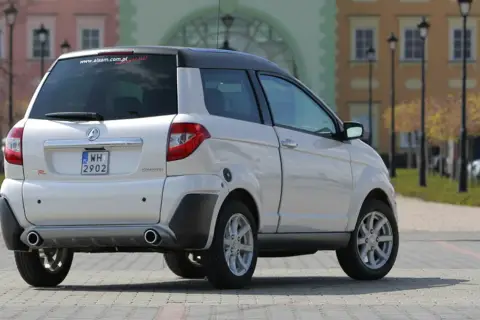 Białe auto Aixam GTR, idealne jako auto bez prawka, parkuje na brukowanym placu przed kolorowymi kamienicami.