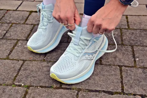 Dłonie zawiązują sznurówki jasnoniebieskich butów do biegania Saucony Ride 18 na chodniku z kostki brukowej.