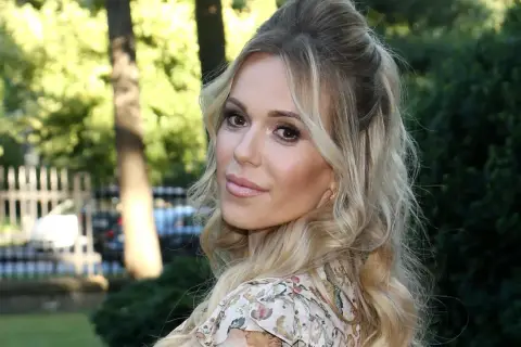 Doda IQ, blondynka w kwiecistej sukience, z burzą loków, uśmiecha się do obiektywu na tle zieleni.