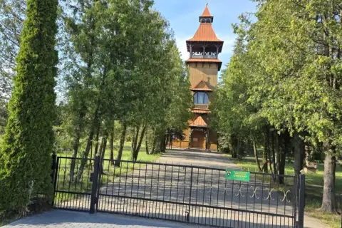 Drewniana wieża kościelna w Długosiodle, otoczona drzewami, za żelazną bramą.