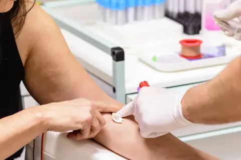 Pobieranie krwi do badań. Ręka pacjentki z widocznymi żyłami, pielęgniarka w białej rękawiczce przygotowuje miejsce wkłucia. W tle probówki i sprzęt medyczny. MPV niskie.