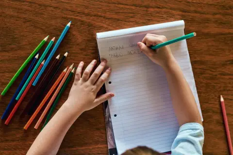 Kind schreibt Namen auf Papier, Teil der schuleingangsuntersuchung was wird gemacht. Buntstifte liegen bereit.