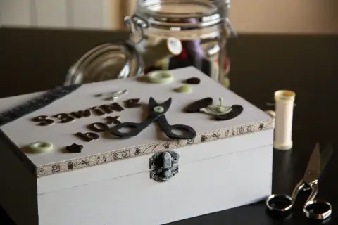 Un costurero pequeño decorado con letras, tijeras de fieltro y botones. Un tarro lleno de hilos y tijeras completan la escena.