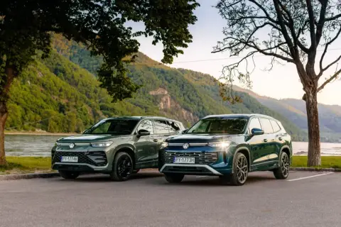 Dwa nowe VW Tayron, zielony i granatowy, zaparkowane nad rzeką, z górami w tle.