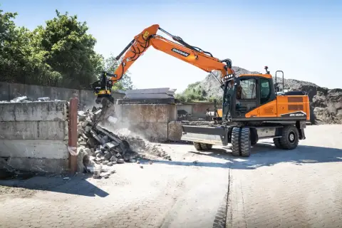 Pomarańczowa koparka Doosan DX150W pracuje na placu budowy w Poznaniu, rozbierając betonowe ściany.