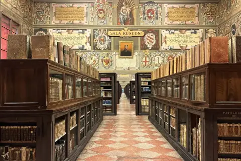 Wnętrze zabytkowej biblioteki z rzędami drewnianych regałów pełnych starych ksiąg, ozdobione malowidłami. To miejsce, gdzie narodziła się nauka, symbolizujące najstarszy uniwersytet na świecie.