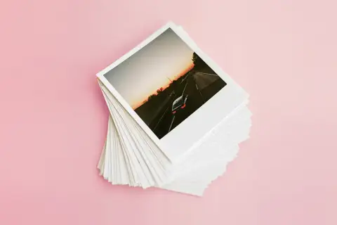 Stos odbitek z wakacyjnej podróży, idealny do wywoływania zdjęć w domu. Na wierzchu zdjęcie samochodu jadącego autostradą o zachodzie słońca.