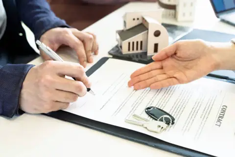 Signature d'un contrat immobilier. Un homme signe un document, des clés et un modèle de maison sont posés sur la table. Le courrier sru est prêt.