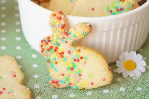 Bunte Osterkekse zum Ausstechen, verziert mit bunten Streuseln, liegen auf einem grünen Tupfentuch. Ein süßer Osterhase steht im Vordergrund.