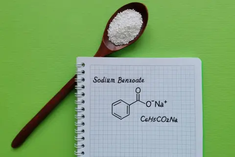 Drewniana łyżka z białym proszkiem, obok notatnik z napisem "Sodium Benzoate" i jego wzorem chemicznym.