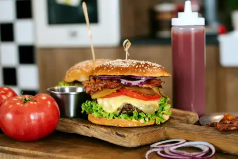 Soczysty burger wołowy z bekonem, serem i warzywami na desce, obok pomidorów i sosu.