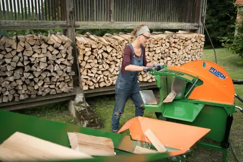 Frau arbeitet mit einer grünen und orangen Wippsäge, um Holzscheite zu schneiden. Im Hintergrund sind Holzstapel zu sehen.