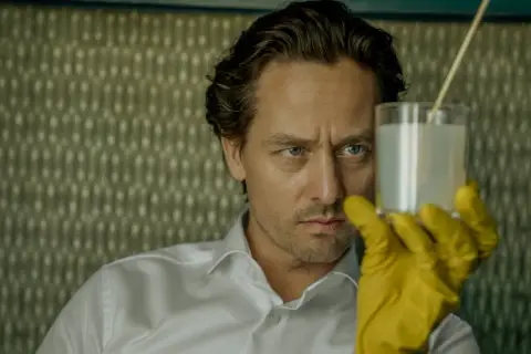 Ein Mann mit blauen Augen und gelben Handschuhen hält ein Glas mit weißer Flüssigkeit. Er ist Teil der "Achtsam Morden Reihe".