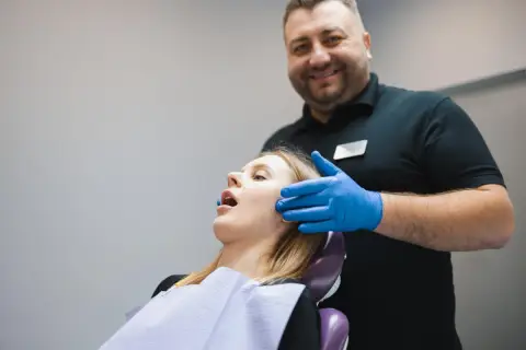 Uśmiechnięty dentysta w niebieskich rękawiczkach bada pacjentkę. Zespół Costena dba o zdrowie jamy ustnej.