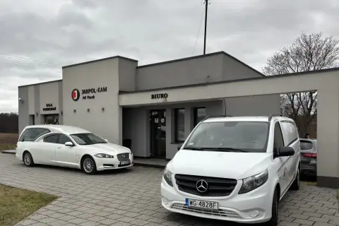 Przed budynkiem "JARPOL-KAM" stoją karawan i van. Firma zajmuje się transportem zmarłych, odpowiadając na pytanie, kto pokrywa koszty sprowadzenia zwłok z zagranicy.