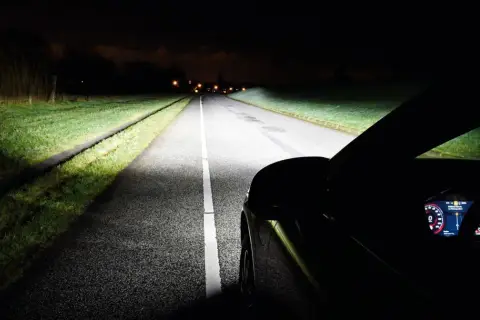 Jasny żarnik reflektora samochodu oświetla nocną drogę, ukazując trawę i zarys asfaltu.