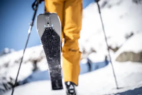 Un skieur en pantalon jaune se prépare à gravir une pente enneigée, sa peau de phoque prête à adhérer à la neige.