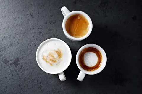 Trzy białe filiżanki z kawą: espresso, latte z wzorem i kawa macchiato na ciemnym tle.