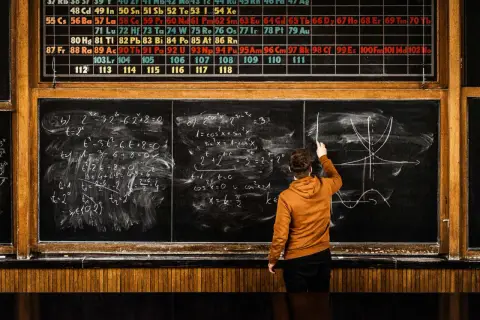 Chłopak na rok przerwy (gap year) rozwiązuje zadania z matematyki i fizyki przy tablicy, nad którą wisi układ okresowy pierwiastków.
