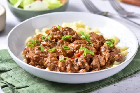 Pyszny przepis na strogonowa z wieprzowiny z pieczarkami i ogórkiem kiszonym, podany z makaronem i posypany szczypiorkiem.