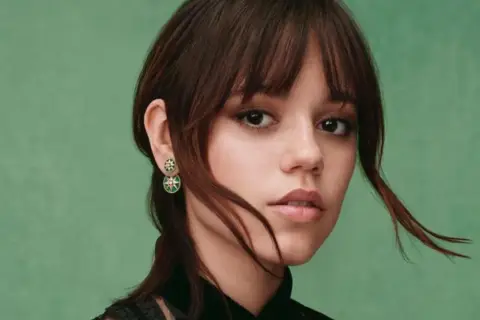 Jenna Ortega z kolczykami w kształcie gwiazdek. Jej włosy są ciemnobrązowe, a tło zielone.