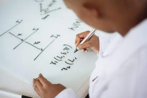 Un niño con bata blanca escribe fórmulas en una pizarra, aprendiendo trucos para resolver problemas de primaria.