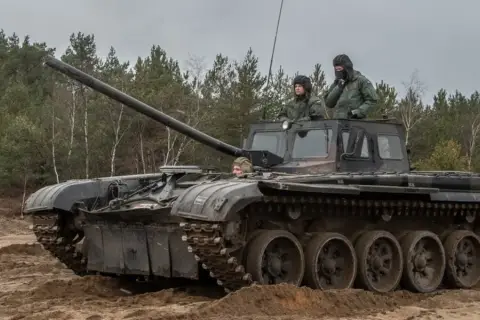 Szkolenie z zakresu jak zostać kierowcą czołgu. Trzech żołnierzy w wojskowych pojazdach, jeden z nich siedzi w otwartym włazie, dwóch innych w kabinie.