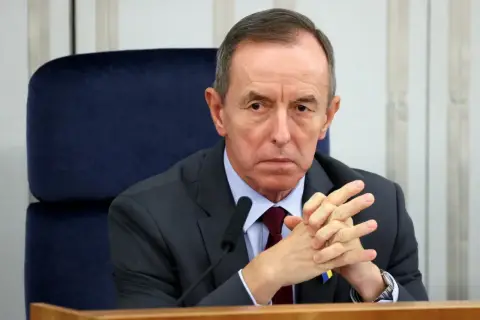 Tomasz Grodzki w garniturze, z rękami złożonymi na piersi, siedzi przy stole. Na klapie marynarki przypinka Poczty Polskiej.