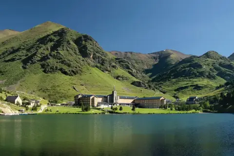 Lago sereno frente a un complejo de edificios en Queralbs, rodeado de montañas verdes y un cielo azul claro.