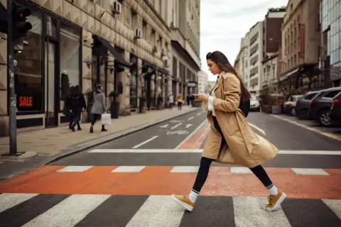 Kobieta w beżowym płaszczu i musztardowych sneakersach przechodzi przez pasy. Idealne buty do trencza na miejski spacer.